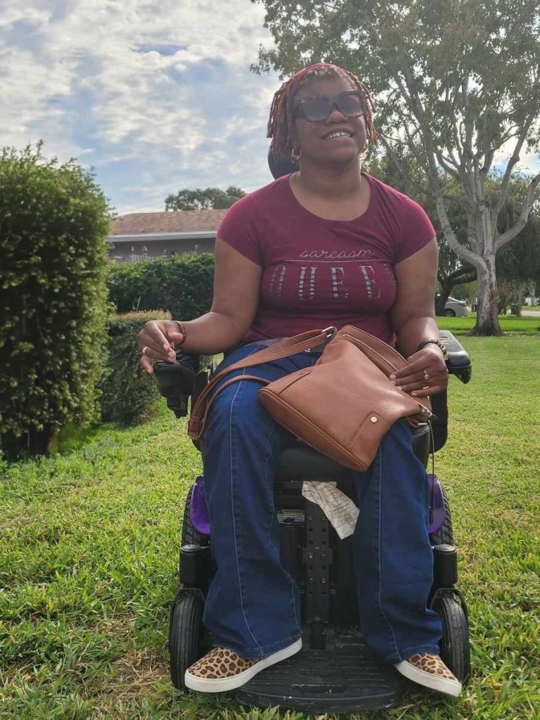 Photo of LeQuina, a black woman with short reddish dreads, sitting in her purple eclectic wheelchair, smiling at the sun.
